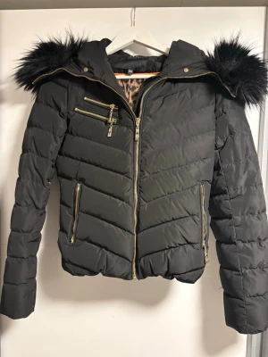 Svart dunjacka med fuskpäls från Hollies - Snygg svart dunjacka från Hollies med guldfärgade dragkedjor, fuskpäls på huvan och leopardmönstrat innerfoder. Jackan har flera fickor med dragkedja och en figurnära passform. Perfekt för kalla dagar när du vill vara både varm och trendig.