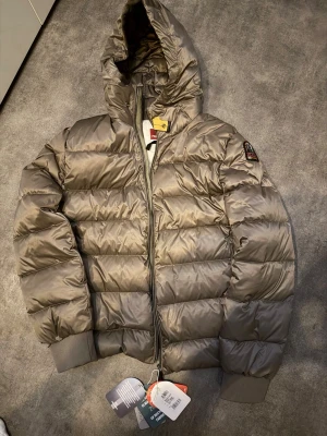 Beige dunjacka från Parajumpers - Snygg beige dunjacka från Parajumpers med huva och quiltad design. Jackan har ribbade muddar vid ärmsluten, dragkedja framtill och klassisk Parajumpers-logga på ärmen. Perfekt för kalla dagar och riktigt stilren look. Passar dig som brukar bära L