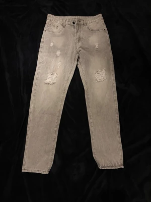 Gråa distressed jeans med raka ben - Säljer ett par beige jeans med raka ben och slitna detaljer framtill. Jeansen har klassisk femficksdesign och knappgylf. Perfekta för dig som gillar en avslappnad och trendig stil.