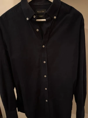 Mörkblå skjorta från Massimo Dutti - Stilren mörkblå skjorta från Massimo Dutti med klassisk krage och vita knappar. Skjortan är långärmad och tillverkad i ett mjukt bomullstyg som känns lyxigt mot huden. Perfekt för dig som gillar en clean och tidlös look.