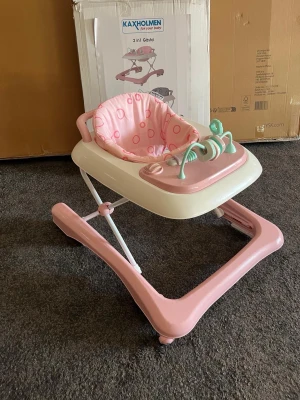 Kaxholmen rosa gåstol med lekbricka - Rosa och vit gåstol från Kaxholmen med mjuk sits i tyg med rosa cirklar. Utrustad med en lekbricka i plast med mintgröna och rosa detaljer, perfekt för små barn. Stommen är i plast och metall, och stolen har hjul för enkel rörelse inomhus.