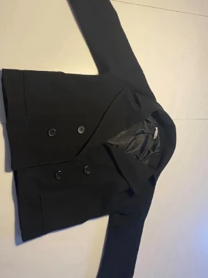 Svart dubbelknäppt kappa  - Stilren svart kavaj med dubbelknäppning och klassisk krage. Kavajen har lång ärm och är fodrad på insidan för extra komfort. Perfekt för en dressad look och enkel att matcha med olika outfits.