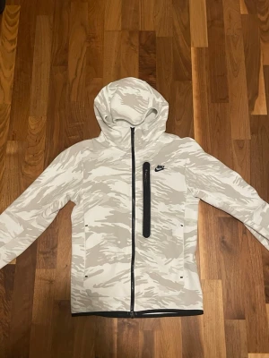 Nike hoodie med beige camomönster - Snygg hoodie från Nike i vitt och beige camomönster. Tröjan har dragkedja framtill, huva och en vertikal bröstficka med svart dragkedja. Perfekt för dig som gillar sportig stil. Materialet känns mjukt och stretchigt. Den är i storlek xs men sitter som en S