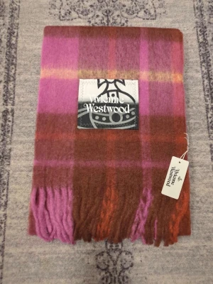 Rutig halsduk från Vivienne Westwood - Snygg rutig halsduk från Vivienne Westwood i lila, rosa, brunt och orange. Mjuk och fluffig känsla med fransar i ändarna. Stor logotyp framtill. Perfekt accessoar för att lyfta din outfit.