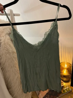 Grön linnetopp med spets - Snygg grön linnetopp med tunna axelband och v-ringning. Toppen har ribbad struktur och fina spetsdetaljer längs urringningen. Fläck på framsidan syns på sista bilden, kanske går bort i tvätten