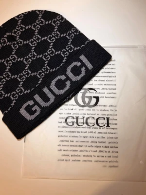 Svart mössa från Gucci med logomönster - Snygg svart mössa från Gucci med grått GG-logomönster och stor GUCCI-text framtill. Klassisk, tight passform och stickad i mjukt material. Perfekt accessoar för att lyfta din streetstyle.