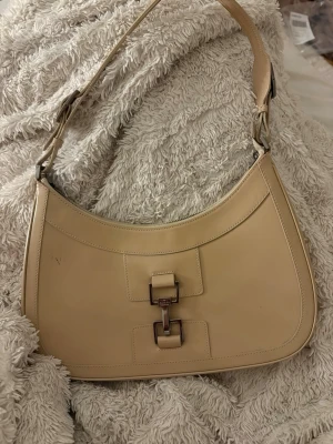 Vintage Gucci horsebit shoulder bag - Säljer en beige axelväska från Gucci i slätt skinn. Väskan har en klassisk halvmåneform, silverfärgade metalldetaljer och ett snyggt spänne framtill. Dragkedja upptill och diskret Gucci-märkning på insidan. Perfekt för dig som gillar stilrena designer. Kan gå ner i pris vid snabb affär 