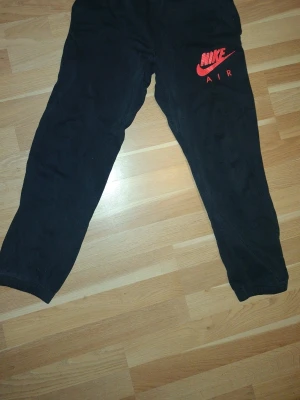 Svarta mjukisbyxor från Nike - Svarta Nike Air mjukisbyxor från Nike med röd logga på benet och liten röd etikett vid bak fickan. Byxorna har normal passform och är gjorda i mjukt bomullsmaterial. Perfekta för chill eller träning, med klassisk Nike-stil.