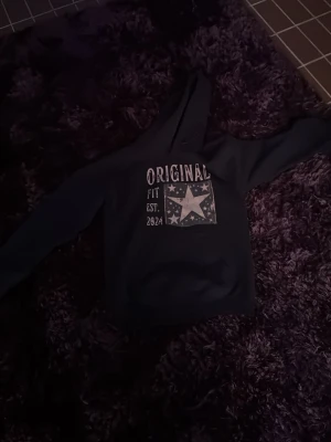 Svart hoodie med tryck och huva - Cool svart hoodie från Original Fit med stor huva och tryck framtill som visar en stjärna och texten 'ORIGINAL FIT EST. 2024'. Hoodien har långa ärmar och en klassisk passform. Perfekt för en avslappnad streetwear-look.