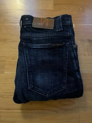 Mörkblå jeans från Nudie Jeans - Säljer ett par mörkblå jeans från Nudie Jeans med klassisk femficksmodell och smal passform. Jeansen har snygga slitningar och kontrastsömmar samt en brun läderpatch med logga bak i midjan. Perfekta för en avslappnad och stilren look.