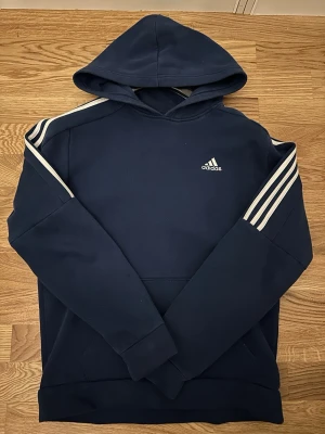 Mörkblå Adidas hoodie med vita ränder - Snygg mörkblå hoodie från Adidas med klassiska vita ränder längs axlarna och ärmarna. Adidas-logga på bröstet och stor huva. Mjuk och bekväm, perfekt för en sportig och avslappnad stil. Känguruficka framtill och ribbade muddar.