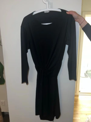 Svart omlottklänning från Filipa K  - En svart omlottklänning från filipa k med lång ärm och knyt i midjan. Klänningen har en enkel sopren design och är tillverkad i ett mjukt, stretchigt material som ger en bekväm passform. Perfekt för dig som gillar stilrena plagg.