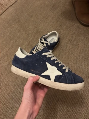 Blåa Golden Goose Superstar sneakers - Säljer ett par mörkblå Golden Goose Superstar sneakers i mocka med vit stjärndetalj på sidan och vita snören. Skorna har en klassisk låg profil och vit sula. Perfekta för dig som gillar en avslappnad men trendig stil.