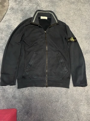 Svart hoodie från Stone Island - Säljer en svart hoodie från Stone Island med dragkedja framtill och två stora fickor. Klassisk patch med logga på vänster ärm och ribbade muddar. Snygg sportig look med diskreta detaljer och hög krage. Perfekt för dig som gillar streetwear.