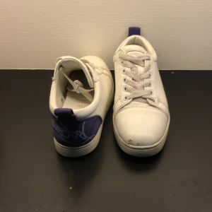 Vita Louboutin sneakers med lila detaljer - Säljer ett par vita sneakers från Christian Louboutin med lila mockadetaljer på hälen och klassisk röd sula. Skorna har snörning, rund tå och är tillverkade i skinn. Perfekta för dig som vill sticka ut med exklusiva och snygga sneakers. Kommer även tvättas när inan ni köper.