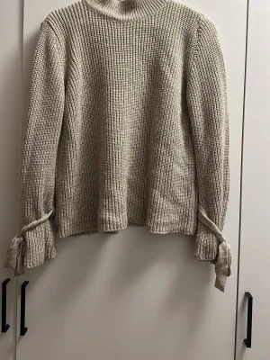 Beige stickad tröja Gina Tricot - Säljer en beige stickad tröja från Gina Tricot med hög krage och långa ärmar. Tröjan har ribbstickad struktur och dekorativa knytband vid ärmsluten som ger en unik touch. Perfekt för dig som gillar en enkel men trendig stil.