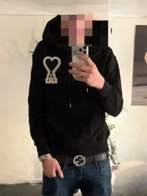 Svart Ami Paris hoodie - Säljer en svart hoodie från Ami Paris med den ikoniska vita hjärtformade A-loggan på bröstet. Perfekt för en avslappnad och trendig stil. Materialet är mjukt bomullstyg.