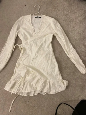 Vit omlottklänning med volang - Snygg vit omlottklänning från Bik Bok med långa ärmar, v-ringning och volangdetalj nertill. Klänningen har knyt i midjan och är tillverkad i ett lätt bomullstyg. Perfekt för en fräsch och stilren look.