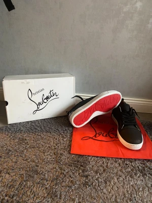 Svarta Christian Louboutin sneakers - Säljer ett par svarta sneakers från Christian Louboutin med klassisk röd sula och vit logga på hälen. Skorna har snörning, vit mellansula och är tillverkade i skinn. Kommer med originalkartong och dustbag.