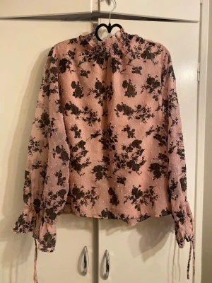 Rosa blommig blus med volangärm - Söt rosa blus med svart blommönster och transparent tyg på baksidan och ärmarna. Blusen har långärmade volangärmar med knytband vid handlederna och smockad halsringning. Perfekt för dig som gillar romantisk och trendig stil. Size is XL but as an M i wear it as oversized blouse.