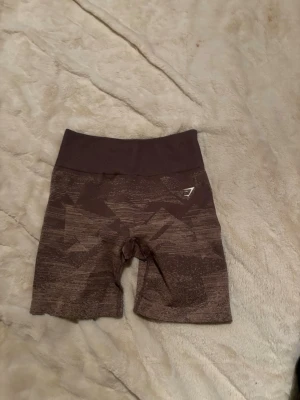 Bruna seamless shorts från Gymshark - Snygga bruna seamless shorts från Gymshark med hög midja och diskret mönster. Stretchigt material som passar perfekt till träning. Logga på vänster sida. Bekväma och stilrena för gymmet eller löprundan.