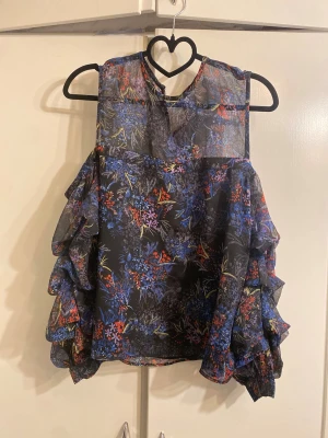 Blommig volangblus med ärmlös design - Snygg blommig blus med volanger längs sidorna och ärmlös design. Toppen har en svart bas med färgstarka blommor i blått, rött och lila. Tillverkad i ett tunt, lätt material som ger ett luftigt intryck. Perfekt för dig som vill sticka ut med en unik topp.