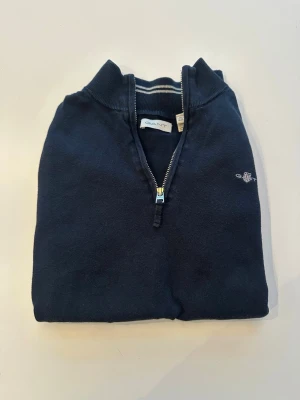 Mörkblå halvzip tröja från Gant - Klassisk mörkblå tröja från Gant med halvzip och ståkrage. Tröjan har ett diskret broderat Gant-logo på bröstet och ribbade muddar vid ärmslut och nederkant. Perfekt för en stilren och avslappnad look.