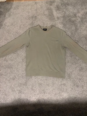Olivgrön sweatshirt från A.P.C. - Snygg olivgrön sweatshirt från A.P.C. med rund halsringning och en bröstficka. Tröjan har en clean look med långa ärmar och ribbade muddar vid ärmslut och nederkant. Perfekt för en avslappnad och stilren outfit.