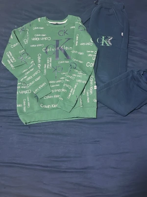 Grön Calvin Klein loungewear set - Snyggt loungewear set från Calvin Klein med en grön sweatshirt och matchande mörkblå byxor. Tröjan har tryckt logga och text i vitt och lila över hela plagget. Byxorna har resår i midjan och logga på benet. Perfekt för chill dagar hemma.