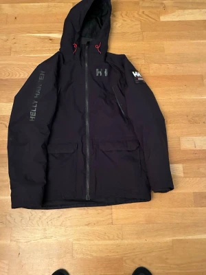 Svart vindjacka från Helly Hansen - Svart vindjacka från Helly Hansen med huva och röda dragskor. Jackan har stora framfickor, dragkedja framtill och logga på bröstet samt ärmen. Perfekt för blåsiga dagar och har en sportig look.