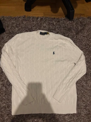 Vit kabelstickad tröja Polo Ralph Lauren - Kabelstickad vit tröja från Polo Ralph Lauren med rund halsringning och den klassiska broderade loggan i marinblått på bröstet. Tröjan har långa ärmar och ribbade muddar vid ärmslut och nederkant. Perfekt för en clean och stilren look.