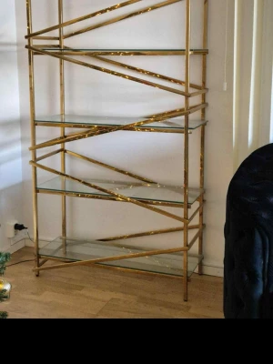 Guldfärgad hylla med glashyllplan - Snygg hylla i metall med guldfärgad ram och fem hyllplan i glas. Hyllan har ett unikt, snedställt mönster på sidorna som ger en modern och lyxig känsla. Perfekt för att visa upp böcker, dekorationer eller växter i vardagsrummet. Jag säljer den på grund av flytt den är cirka 2 år gammal 
