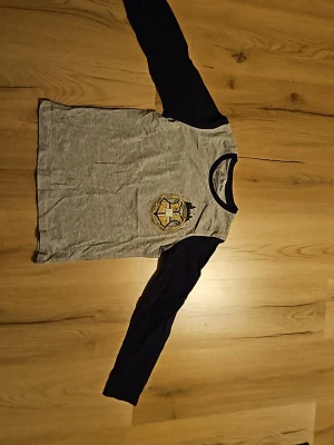 Harry Potter långärmad t-shirt - Mysig långärmad pyjamaströja från Harry Potter med grå kropp och svarta ärmar. Tryck med Hogwarts-emblem på bröstet och kontrastfärgad svart halsringning. Perfekt för alla Potterheads som vill chilla i stil. Använd 2 gånger.