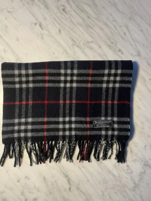 Berberry halsduk 100% kashmir  - Säljer en klassisk rutig halsduk från Burberrys of London i 100% mjuk kashmir. Den har en snygg svart bas med vita och röda rutor samt traditionella tvinnade fransar i ändarna. Halsduken är märkt med Burberrys of London