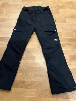 Svarta skidbyxor från WARP - Svarta skidbyxor från WARP med raka ben och praktiska fickor fram och bak. Byxorna har justerbar midja och reflexdetaljer på baksidan av benen. Perfekta för kalla vinterdagar och skidåkning.