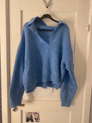 Blå stickad tröja med krage - Mysig blå stickad tröja med v-ringning. Tröjan har en avslappnad passform, långa ärmar och ribbstickat material. 