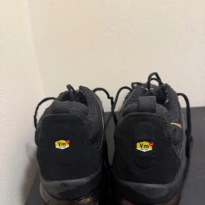 Nike Air Vapormax svarta/guld sneakers - Svarta Nike Air Vapormax sneakers med guldfärgade detaljer och synlig luftdämpning i sulan. Skorna har snörning, rund tå och en cool, futuristisk design med plastdetaljer längs sidorna. Ovandelen är i textil och syntet, perfekt för dig som gillar streetwear.