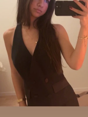 Svart  med v-ringning - Säljer en svart byxdress med djup v-ringning och dubbla knappar framtill. Byxdressen har en elegant och stilren look med fickdetaljer och är ärmlös. Perfekt för dig som vill ha en dressad och trendig outfit.