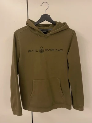 Sail Racing hoodie - Olivgrön hoodie från Sail Racing med svart logga tryckt på bröstet och en mindre logga på huvan. Klassiskt mjukt material som känns skönt mot huden. Perfekt för en avslappnad och sportig stil.