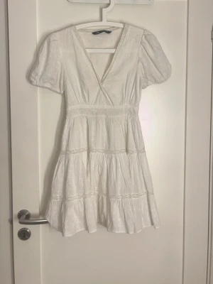 Vit omlottklänning med puffärm - Kort vit klänning från ZARA med omlott-v-ringning, puffärmar och volangdetaljer. Tillverkad i lätt bomullstyg med diskret broderat mönster och spetsinslag. Perfekt för sommarens alla tillfällen.använd endast 1 gång