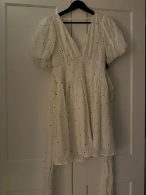 Vit prickig omlottklänning med puffärm - Supersöt vit kortklänning med svarta prickar och puffiga korta ärmar. Klänningen har omlottmodell med v-ringning och knytband i midjan. Tillverkad i ett lätt och luftigt material, perfekt för varma dagar. Oanvänd kvar med prislapp! Storlek 34 