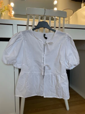 Vit blus med puffärm och knyt - Söt vit blus med puffiga korta ärmar och två knytband framtill. Blusen har en avslappnad passform och lätt rynkad nederdel. Tillverkad i ett tunt, vävt bomullstyg som känns luftigt och fräscht. Perfekt för en trendig och enkel look.
