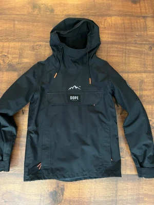 Svart skidjacka från Dope Snow - Svart vindjacka från Dope Snow med stor huva, snörning och en stor magficka med DOPE-logga. Jackan har orange detaljer på snören. Tillverkad i slitstark polyester och har lång ärm. Finns fickor för liftkort på armen!