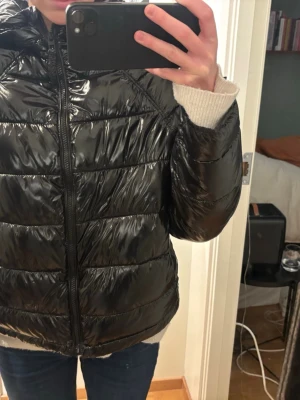 Svart glansig pufferjacka med huva - Säljer en svart, glansig pufferjacka med huva och dragkedja framtill. Jackan har quiltad design och lång ärm, perfekt för kyliga dagar. Passformen är normal och materialet känns som syntet. Enkel och stilren look som funkar till det mesta.