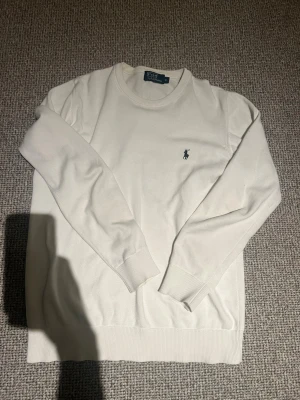 Vit tröja från Polo Ralph Lauren - Klassisk vit tröja från Polo Ralph Lauren med rund halsringning och broderad marinblå logga på bröstet. Tröjan har långa ärmar och ribbade muddar vid ärmslut och nederkant. Perfekt för en clean och stilren look. Tröjan är i M och skulle säga att den sitter som bäst mellan 180-185 
