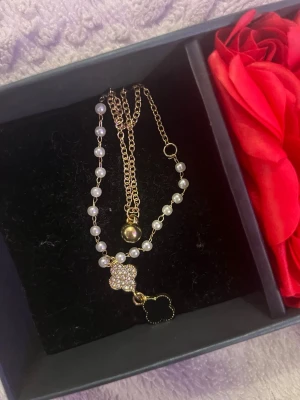 Guldhalsband med pärlor och klöverhänge - Snyggt halsband med guldfärgad kedja och vita pärlor. Hängsmycket består av en glittrande klöverformad detalj med stenar och en svart klöver. Perfekt accessoar för att lyfta din outfit med en trendig och elegant touch.