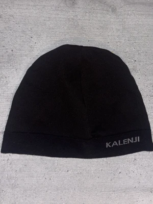 Svart mössa från Kalenji - En stilren svart mössa från Kalenji med diskret logga framtill. Tillverkad i ett tunt, stretchigt material som passar perfekt för löpning eller andra utomhusaktiviteter. Enkel och sportig design som sitter skönt på huvudet.