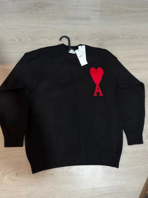  Ami Paris - Svart stickad tröja från Ami Paris med röd hjärtlogga och bokstaven A på bröstet. Tröjan har rund halsringning, ribbade muddar och nederkant. Perfekt för dig som gillar stilrena plagg med en twist.