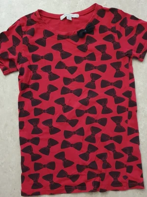 T-shirt - Cool röd t-shirt från Mywear Young med svarta rosetter över hela plagget och en extra tygrosett vid halsen. T-shirten har rund halsringning och korta ärmar. Perfekt för dig som vill sticka ut med ett lekfullt mönster och skön passform.