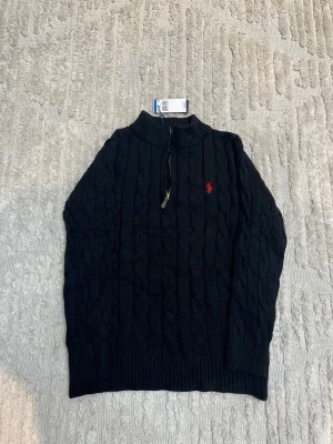 Svart stickad tröja från Polo Ralph Lauren - Kabelstickad svart tröja från Polo Ralph Lauren med halv dragkedja och röd broderad logga på bröstet. Tröjan har hög krage och långa ärmar, perfekt för kyliga dagar. Klassisk och stilren design som funkar till det mesta.